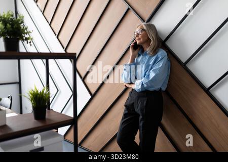 Fifty year old successful business woman executive talking on mobile phone leaning against the wall in the office. Professional Stock Photo