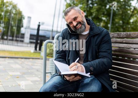 A man in a city park sits on a bench I am talking on the phone writes in a notebook Stock Photo