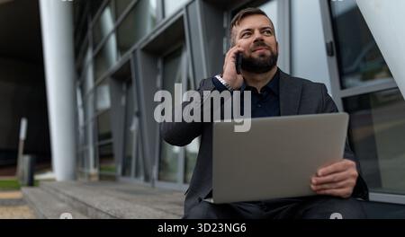 Businessman talking on a mobile phone with a laptop on his knees while sitting at the entrance to the office building Stock Photo