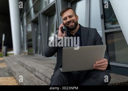 Business analyst in a strict business suit talking on a mobile phone and working on a laptop next to a business center Stock Photo