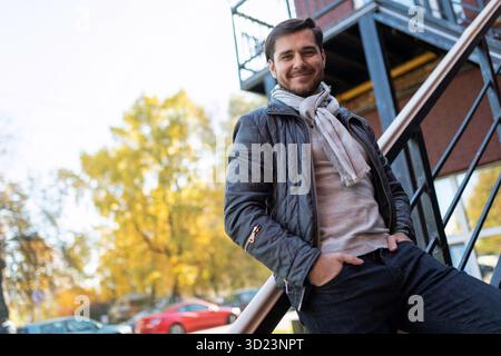 Pretty with a stylish young man stands leaning on the stairs against the backdrop of the autumn city Stock Photo