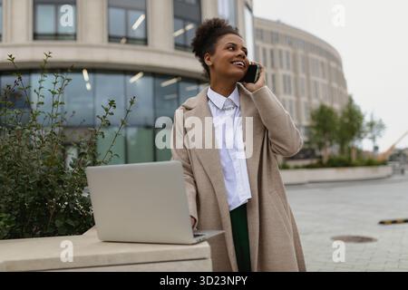 African american woman entrepreneur working on a laptop online and talking on a mobile phone with a smile on the background of a Stock Photo