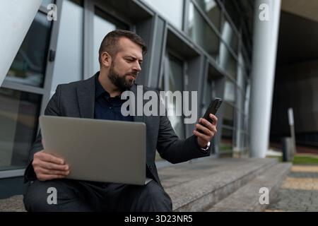 A man works with a laptop online and watches a mobile phone while sitting near the office building, online banking concept Stock Photo