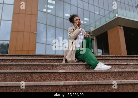 African american business woman talking on a mobile phone while sitting on the steps in front of an office building Stock Photo
