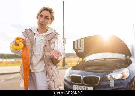 Upset woman near a broken car with a tow rope in her hands, the concept of life and vehicle insurance against accidents Stock Photo