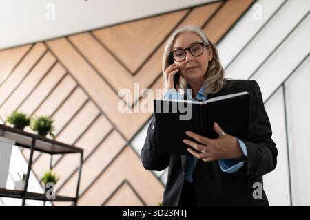 Mature businesswoman in glasses and business clothes speaks on a mobile phone with a notepad in her hands in the office Stock Photo