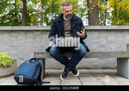 Male businessman on a business trip with a backpack working on a laptop while sitting in a city park, business risk insurance co Stock Photo