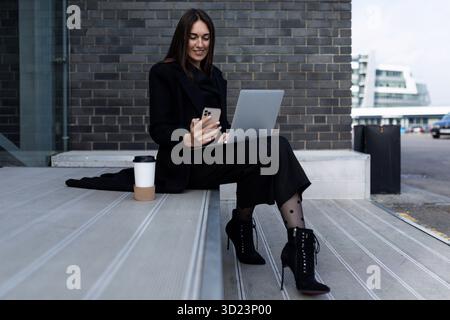 Business modern independent woman working on a laptop with a phone in her hands outside the office Stock Photo