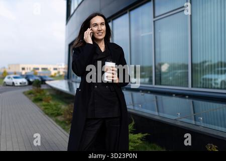 A woman specialist in dealership goes to the office talking on a mobile phone with a glass of coffee in her hands, Supporting cu Stock Photo