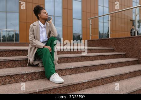 Young american female freelance marketer talking on the phone while sitting on the steps of an office building Stock Photo
