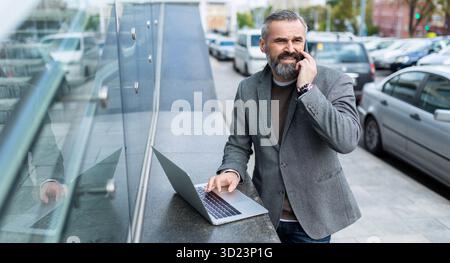 A businessman in a gray jacket works online on a laptop next to an office building and speaks on a mobile phone with a smile Stock Photo