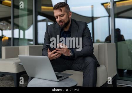 A man in a business suit works in a street cafe on a laptop with a mobile phone in his hands and smokes a cigarette Stock Photo