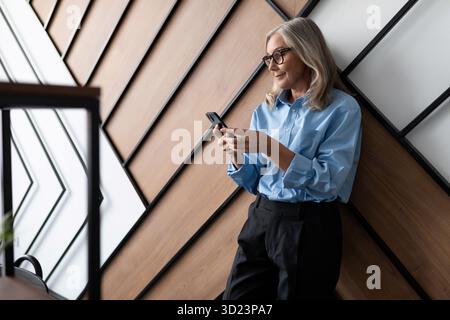 Adult mature independent successful business woman in a blue shirt with a phone in her hands on the background of a modern offic Stock Photo
