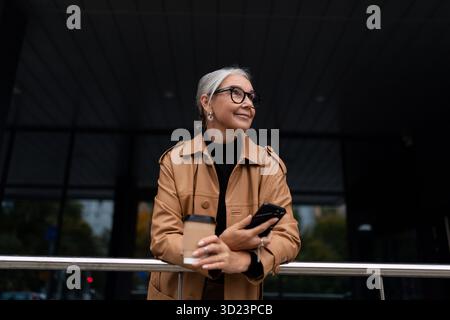 An elderly mature woman on the phone and a cup of coffee in her hands looks into the distance outside Stock Photo