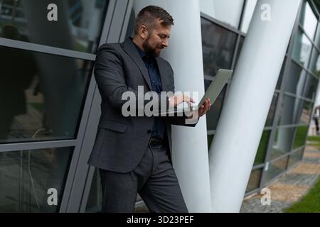 A man in a business suit is typing on a laptop standing next to an office building Stock Photo