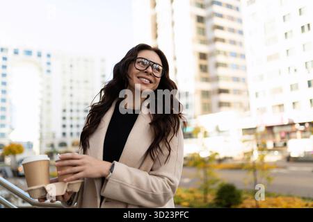 Stylish and satisfied businesswoman with cups of coffee in her hands on the background of the urban landscape Stock Photo