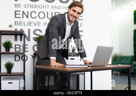A charismatic businessman in a business suit stands leaning over his desk and looks at the camera Stock Photo