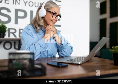 Woman manager 50 years old sits at a table in headphones during an online conference looks into a laptop. . Tax accountant Consu Stock Photo