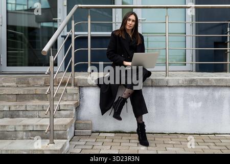 Successful business woman working on laptop online sitting on stairs wearing stylish black coat Stock Photo