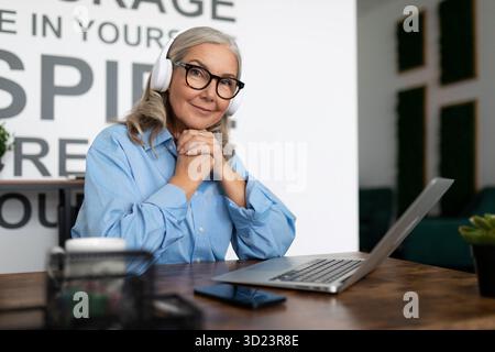 Successful mature woman manager watching training video on laptop with headphones sitting at table in office. Accounting Bookkee Stock Photo