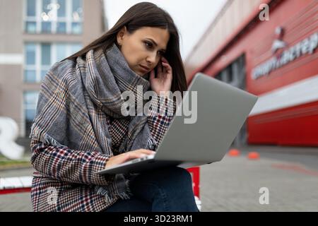 Young serious woman businesswoman focused working on laptops against the backdrop of the city in warm autumn clothes Stock Photo