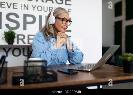 Business serious mature businesswoman watching a webinar on a laptop in the office with her hands folded under her chin in headp Stock Photo