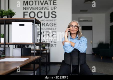 Adult woman executive talking on a mobile phone while sitting on a chair backwards in the office. Bank Advisor And Auditor conce Stock Photo