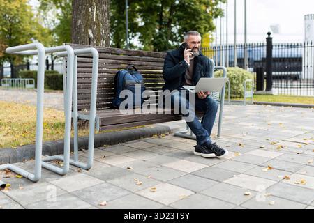 Adult male businessman working online on laptop and calling by phone in city park Stock Photo