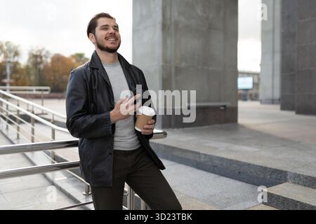 Young man with a cup of coffee by a mobile phone with a wide smile on his face next to a city building Stock Photo