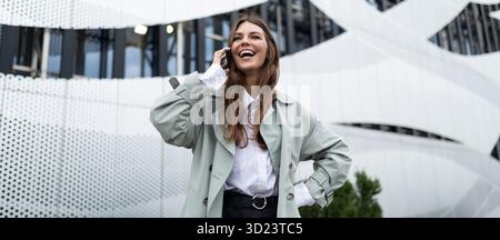 A young woman speaks on a mobile phone with a wide smile on her face against the background of an aluminum facade of an office b Stock Photo