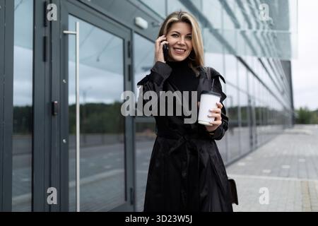 Happy joyful smiling caucasian woman in black business clothes talking on the phone with a cup of coffee in her hands outside Stock Photo