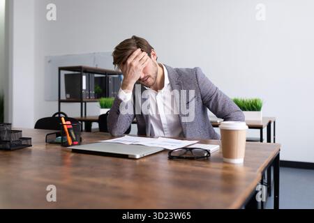 Tired upset businessman sits in front of the desktop in the office covering his face with his palm Stock Photo
