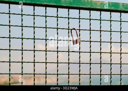 A rusted padlock hangs on a green wire fence with a blurred urban skyline in the background. Stock Photo