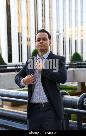 Confident latin businessman in formal attire adjusts his suit jacket while standing outside near modern office buildings Stock Photo