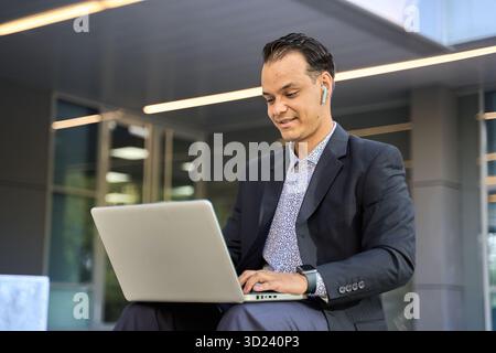 Latin businessman in suit using laptop with wireless earphones, working remotely outside a modern glass building Stock Photo