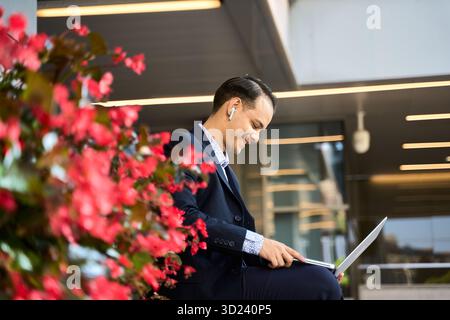 Smiling latin businessman using laptop and earphones outdoors, sitting by vibrant flowers in a modern urban office setting Stock Photo