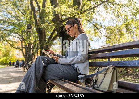 Focused businesswoman reading on tablet at park bench remote work outdoors with deep concentration, analyzing digital documents and market strategy, c Stock Photo