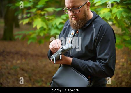 A focused man adjusts his drone while surrounded by vibrant trees displaying shades of green and gold. It is a peaceful afternoon in nature. Stock Photo