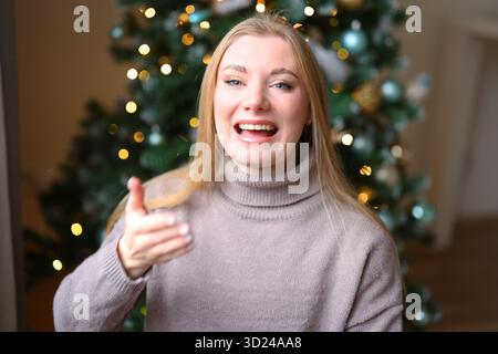 Smiling blonde woman in sweater with glowing Christmas lights behind, celebrating joyful holiday season. Stock Photo