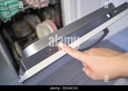 Female hand pressing start button on dishwasher door with clean dishes inside Stock Photo