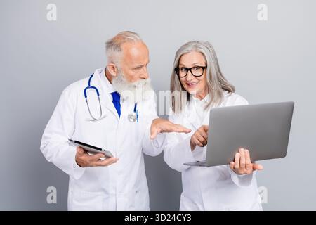 Two experienced doctors review patient data together in a clinic using tablet and laptop and discuss diagnosis and treatment Stock Photo