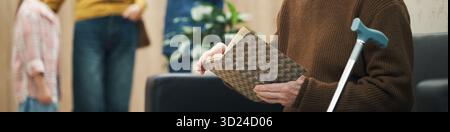 Header of senior man sitting with walking cane holding book, reading while waiting in queue in medical clinic reception area for doctor appointment Stock Photo