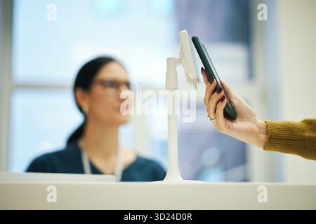 Anonymous woman holding smartphone making contactless payment at checkout counter at clinic reception, while young female nurse in medical uniform with glasses observing transaction process Stock Photo
