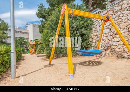 Empty blue basket swing on a yellow swing set in a sunny, fenced playground with a sandy ground, green bushes, and stone wall Stock Photo