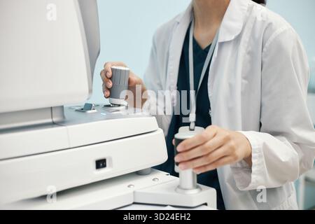 Young female doctor in white coat operating ophthalmology diagnostic equipment in clinical setting, hands adjusting controls on advanced machine, wearing lab coat and identification badge Stock Photo