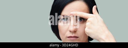 Caucasian young female with black hair pointing to forehead wrinkle on pale Stock Photo