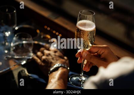 Close-up of a man's hand holding a glass of sparkling wine or champagne during sunset with reflections on glass table. Stock Photo