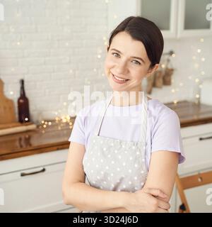 Smiling young caucasian female chef in polka dot apron posing in bright kitchen Stock Photo