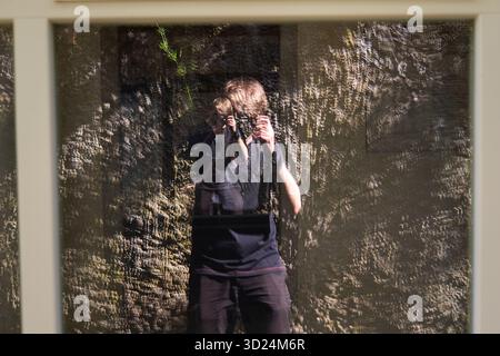 Reflection of a photographer taking a picture through a glass window with a textured stone wall behind. Stock Photo