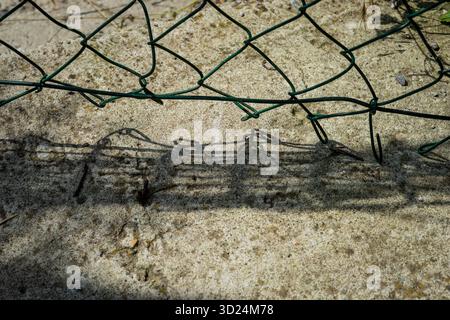 Broken green metal fence casting a geometric shadow on rough concrete surface in sunlight. Stock Photo